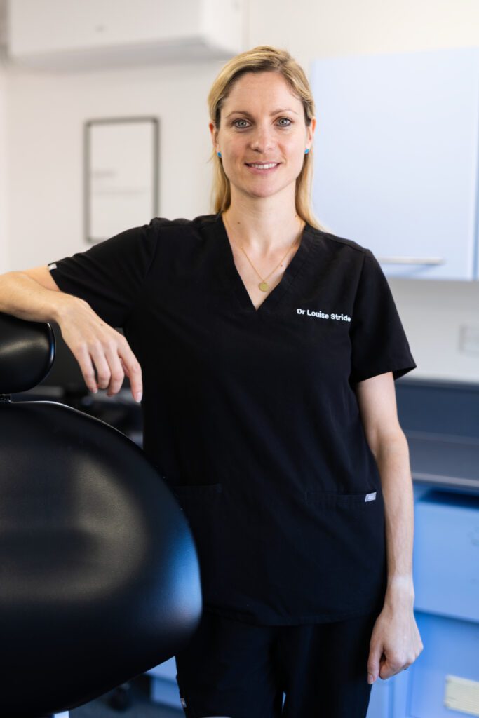 Dr Louise Stride standing in a dental surgery, smiling and wearing black clinical uniform beside a dental chair