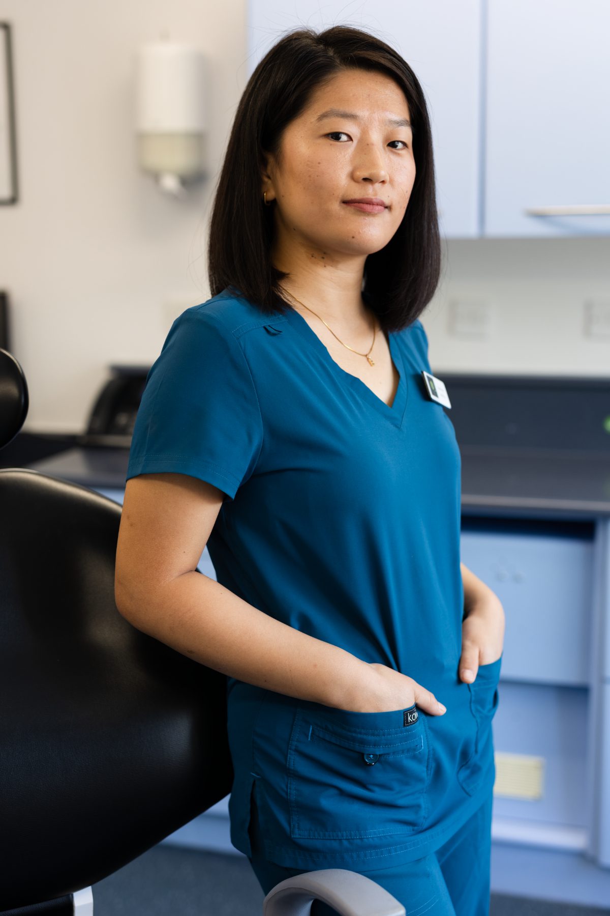 Dental nurse Buduna Chemjong Limbu standing in a dental surgery, wearing teal clinical scrubs with hands in pockets beside a dental chair.
