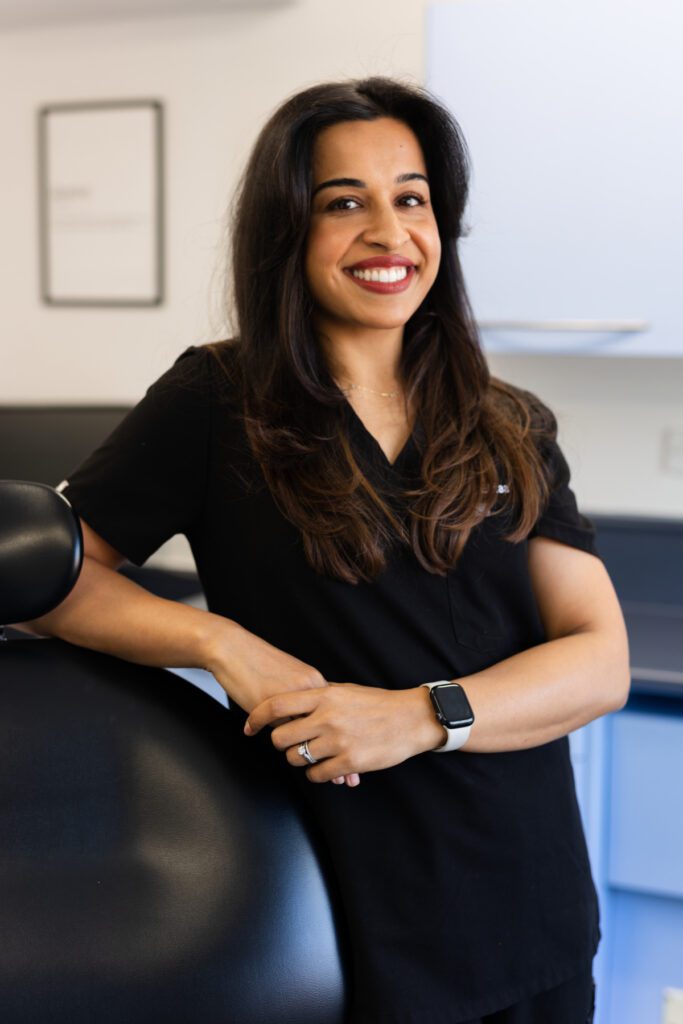 Female dental professional Ektaa Chopra smiling in a dental surgery, wearing black clinical uniform and resting her arm on a dental chair.