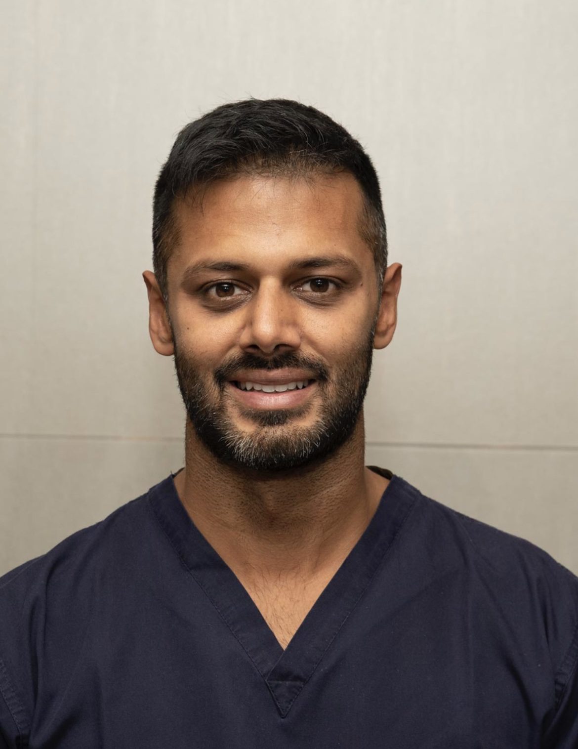 Dentist Noman Athwal smiling against a neutral background, wearing dark clinical scrubs.