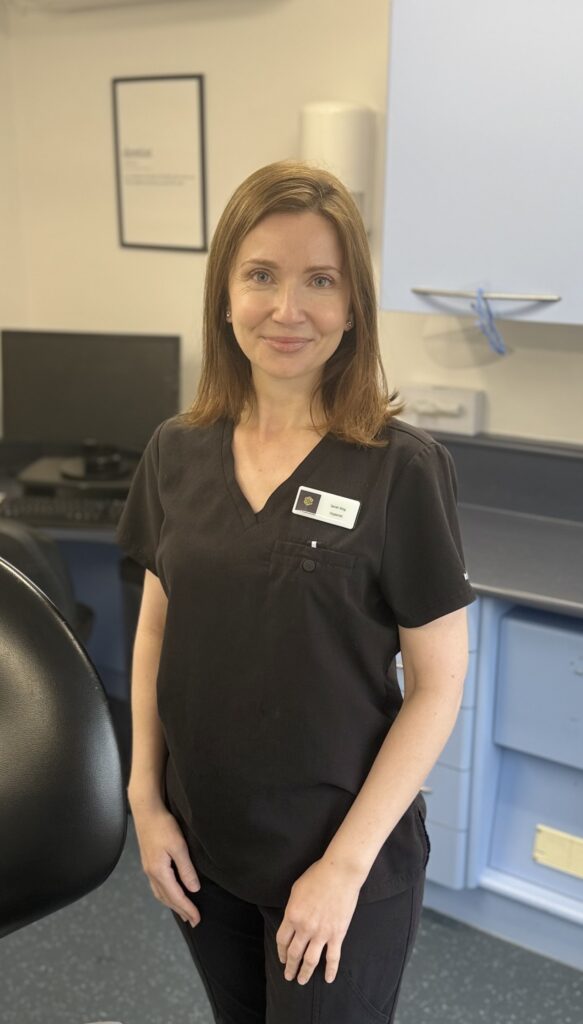 Dental hygienist Sarah King smiling in a dental surgery, wearing black clinical uniform and standing beside a dental chair.
