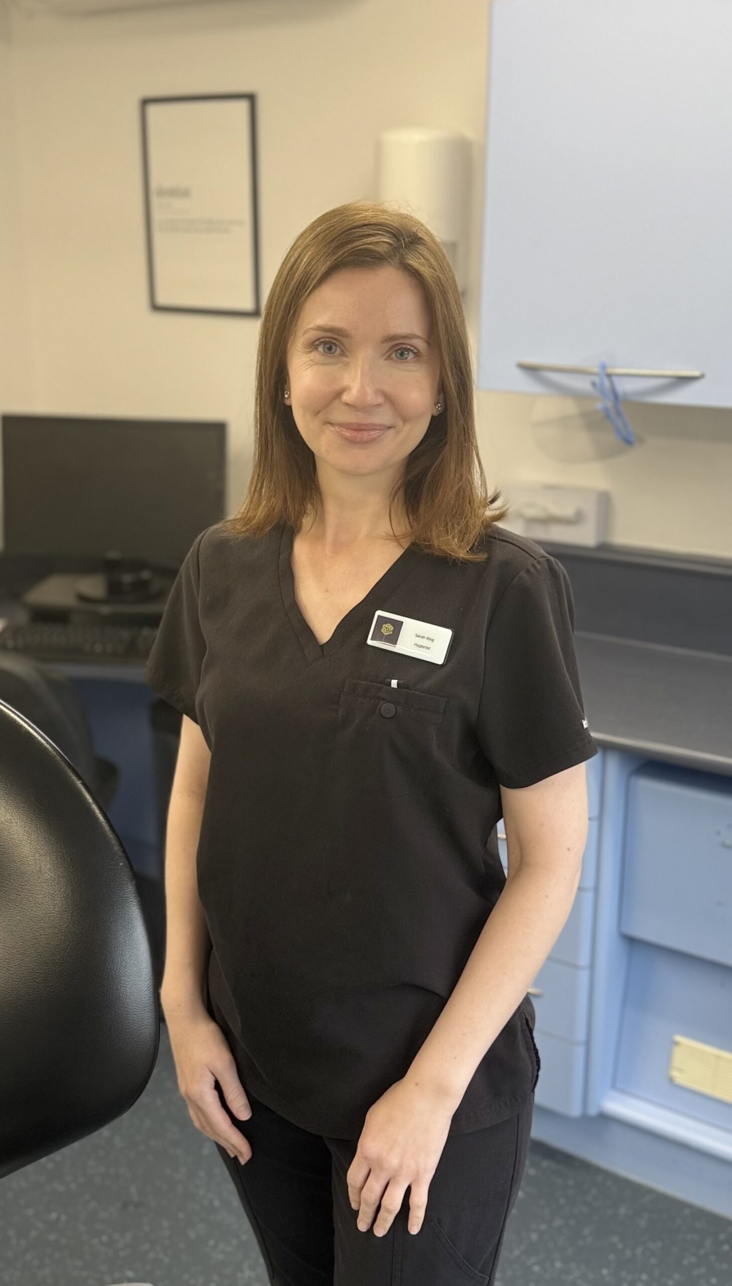 Dental hygienist Sarah King smiling in a dental surgery, wearing black clinical uniform and standing beside a dental chair.