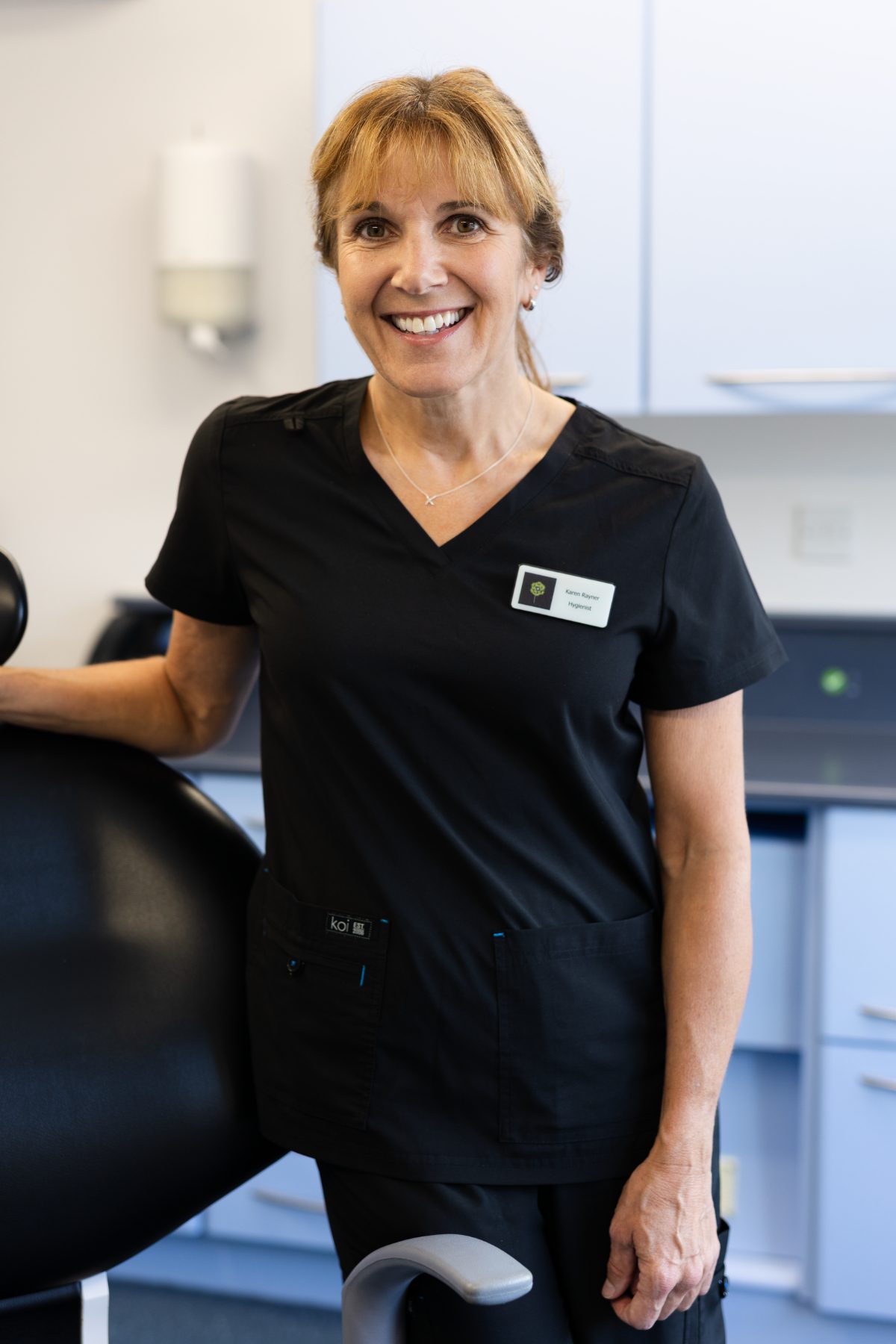 Dental hygienist Karen Rayner smiling in a dental surgery, wearing black clinical uniform and resting her hand on a dental chair.