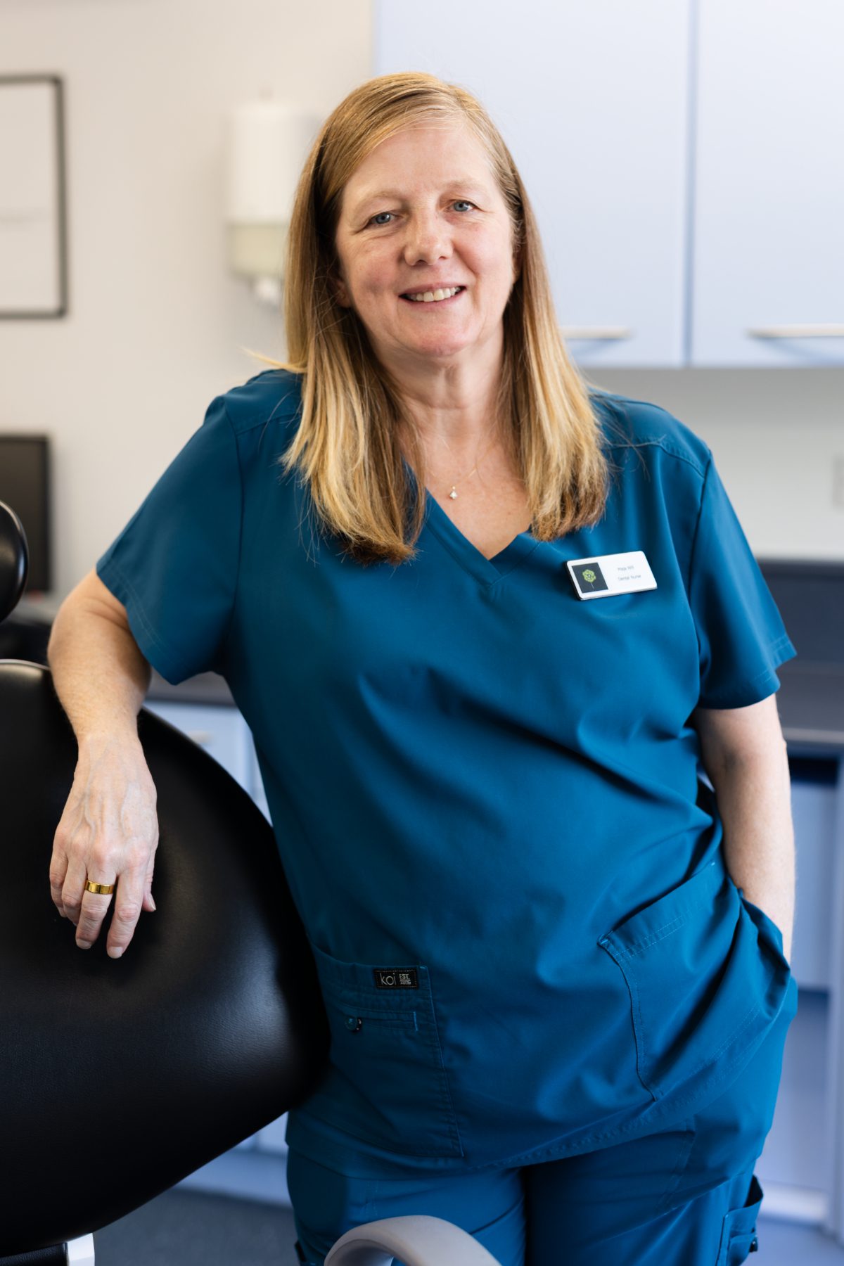 Dental nurse Maja Will smiling in a dental surgery, wearing teal clinical scrubs and resting her hand on a dental chair.