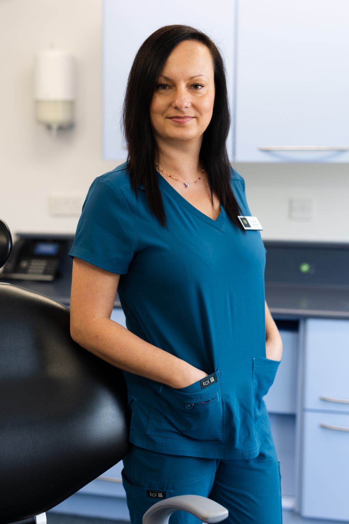 Dental nurse Maria Kujawa smiling in a dental surgery, wearing teal clinical scrubs with hands in pockets beside a dental chair.