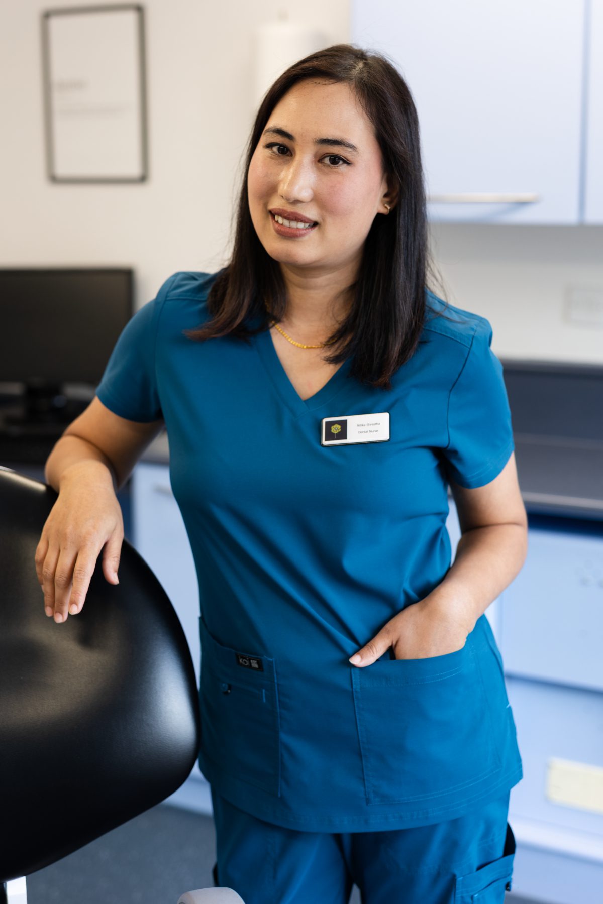 Dental nurse Nitika Shrestha smiling in a dental surgery, wearing teal clinical scrubs and resting her hand on a dental chair.