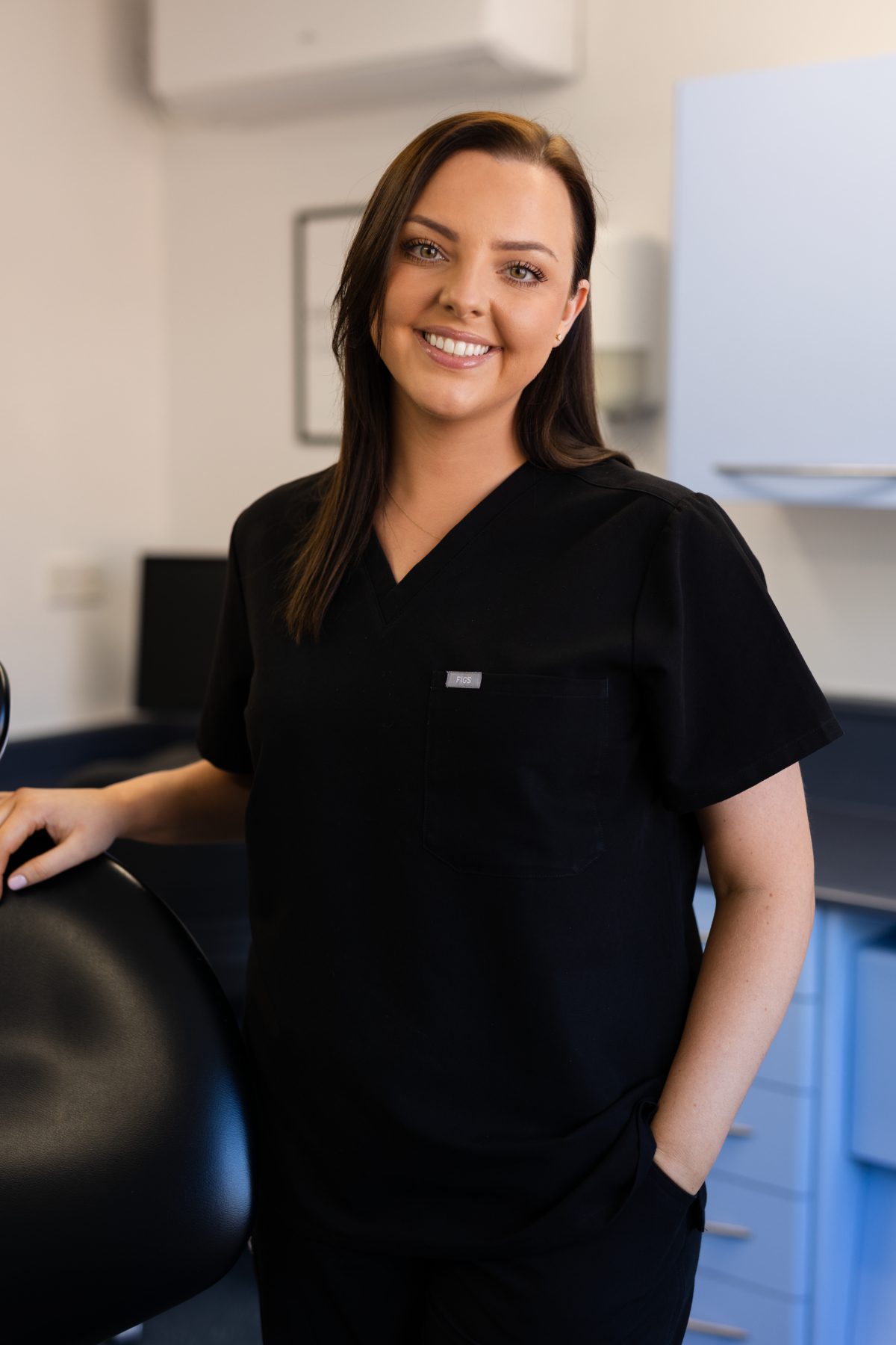 Dental hygienist Sarah Pritchard smiling in a dental surgery, wearing black clinical uniform and resting a hand on a dental chair.