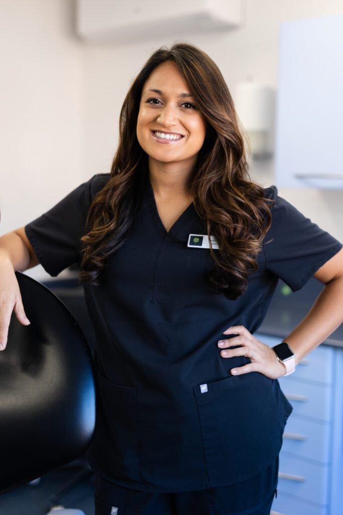 Shreena Patel smiling in a dental surgery, wearing black clinical uniform and standing beside a dental chair.