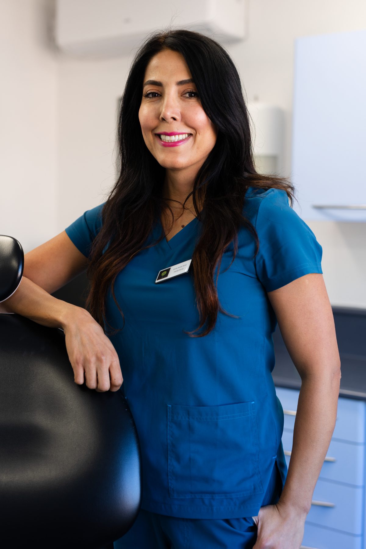 Trainee dental nurse Sonia Amiri smiling in a dental surgery, wearing teal clinical scrubs and resting her hand on a dental chair.