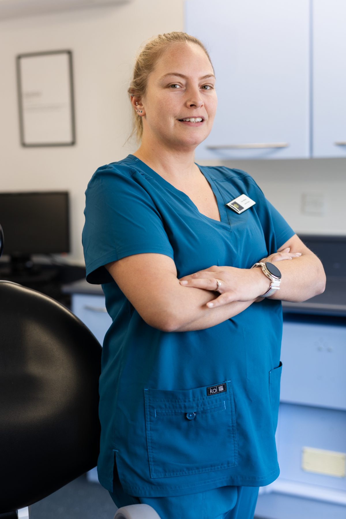 Senior dental nurse Vicki Williams smiling in a dental surgery, wearing teal clinical scrubs with arms folded beside a dental chair.