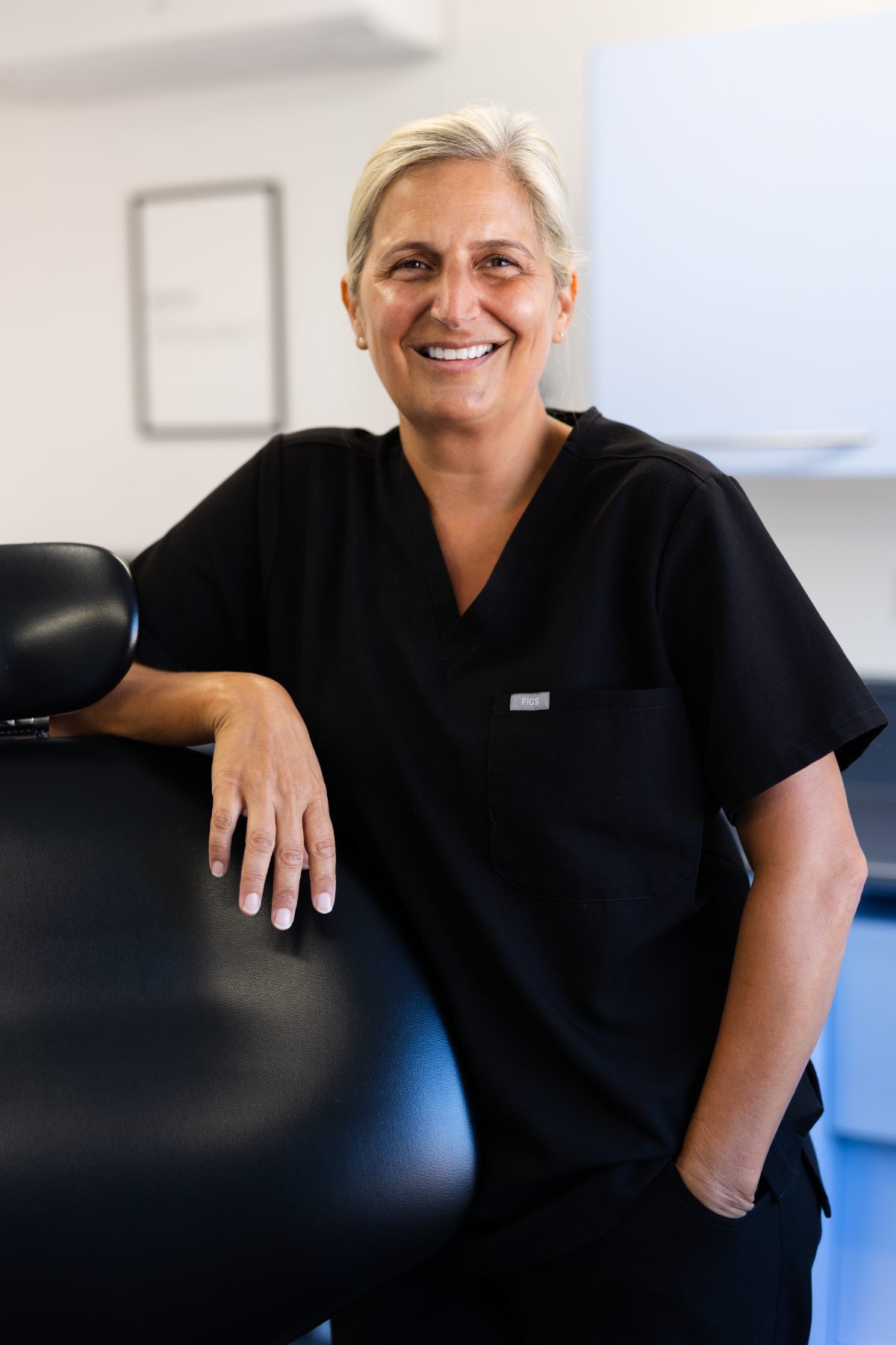 Dental hygienist Victoria Bonilla smiling in a dental surgery, wearing black clinical uniform and resting her hand on a dental chair.