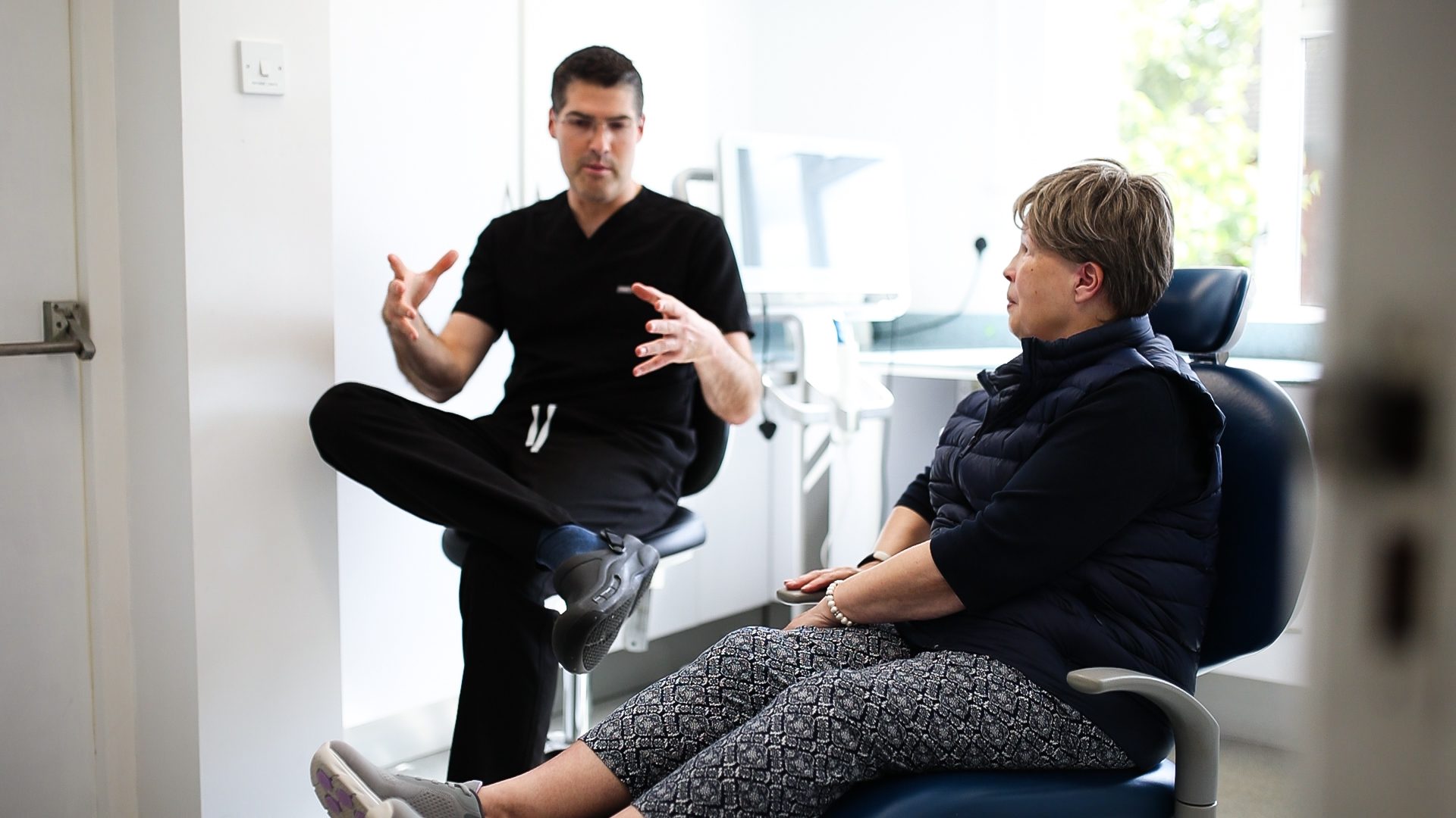 Male dentist sitting in a relaxed pose explaining a treatment to a female patient sat in the dentist chair