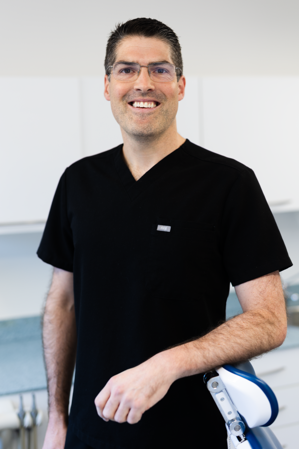 Ben Kemsley, Principal Dentist, smiling and standing in a dental surgery wearing black clinical uniform.